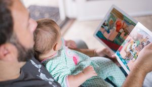 A father seated in an armchair reads a picture book to the baby on his lap