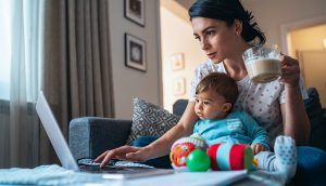 A mother holds her coffee as she works on a laptop with a baby seated on her lap