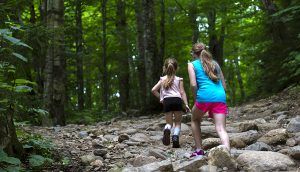 An older and younger girl are running through a rocky clearing in the woods.