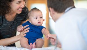 A mother holds her baby sitting up on an examination table as a doctor and holds the baby's hand.