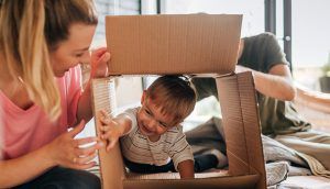A smiling boy climbs through an open cardboard box lying on its side as a man and woman look on.