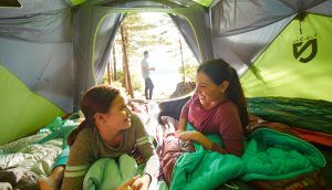 A woman and girl smile and laugh together in a tent as a man can be seen going for a walk outside.