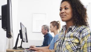 A woman is seated at a computer, smiling as she views the screen