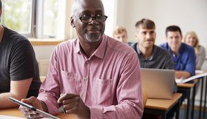 An older man is seated at the front of a class of adult learners, holding a clipboard
