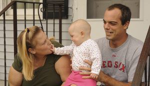 A seated man holds up a baby, who reaches an arm out to a seated woman who kisses her hand.