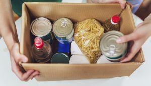 Hands can be seen holding a cardboard box, with an open top revealing bottles and cans of food.