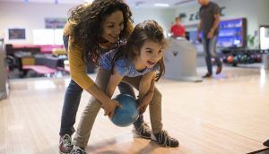 In a bowling alley, a woman helps a young girl roll a ball down the lane.