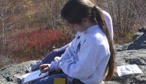 Girl wearing a sweatshirt sits on a rock outside coloring on a clipboard