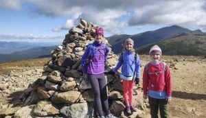 Three children stand smiling in front of a pile of stones outdoors, with mountains visible in the background.