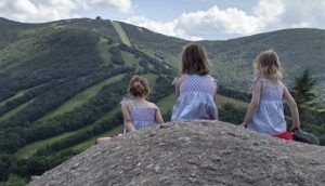 Three girls are seated on a small hill, looking out at the mountains